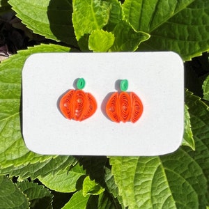May include: A pair of orange pumpkin-shaped earrings with green stems. The earrings are made of paper and are displayed on a white card.