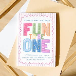 Könnte beinhalten: Eine farbenfrohe Geburtstagseinladung mit dem Text "FUN TO BE ONE" in Buchstaben. Die Einladung befindet sich auf einem cremefarbenen Hintergrund, mit einem Holzrahmen und einem weißen Papier. Die Einladung enthält Datum, Uhrzeit und Ort.