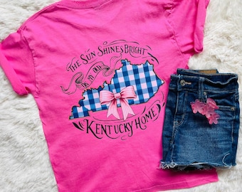May include: Pink t-shirt with a blue and white gingham Kentucky state outline and a pink bow. The text "The Sun Shines Bright On My Old Kentucky Home" is printed on the shirt.