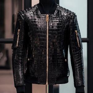 May include: A black jacket with a glossy crocodile skin texture, featuring a prominent gold-toned central zipper and matching gold-toned zippers on each sleeve. The garment includes a black ribbed collar, cuffs, and hem, displayed on a mannequin.
