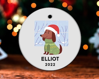 May include: A white, round Christmas ornament featuring a cartoon dachshund wearing a green sweater and Santa hat, with a snowy window background. The name "ELLIOT" and the year "2022" are printed below the dog.