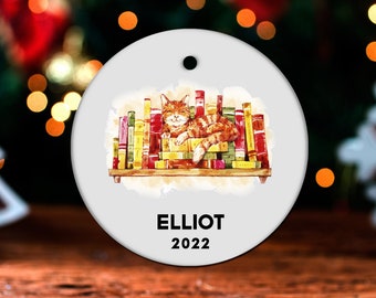 May include: A round white Christmas ornament with a watercolor illustration of a cat sleeping on a stack of books. The ornament is personalized with the name "ELLIOT" and the year "2022".