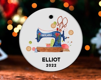 May include: A white round Christmas ornament with a blue sewing machine, red thread, and colorful buttons. The ornament has the text "HANDMADE" on the sewing machine and "ELLIOT 2022" at the bottom.