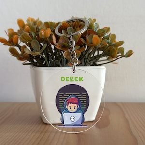 Könnte beinhalten: Ein runder, transparenter Schlüsselanhänger aus Acryl mit einer Cartoon-Illustration einer Person, die ein grünes Kapuzenoberteil trägt und einen Laptop benutzt. Der Laptop hat ein Totenkopf-Motiv. Der Text "DEREK" ist in grünen Buchstaben auf dem Schlüsselanhänger gedruckt.