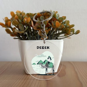 May include: A clear round keychain with a white background and a cartoon illustration of a person riding a camel in front of the pyramids. The text "DEREK" is printed in black at the top of the keychain.