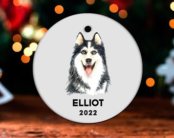 May include: Round white ceramic ornament with a watercolor painting of a black and white husky dog. The dog is looking at the camera with its tongue sticking out. The ornament has the name "ELLIOT" and the year "2022" printed on it.