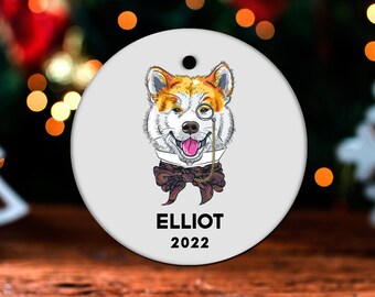 May include: White round Christmas ornament with a cartoon illustration of a dog wearing a bow tie and a monocle. The dog has orange and white fur. The text "ELLIOT 2022" is printed below the dog.