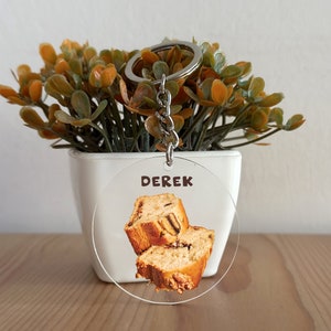 Op de afbeelding: Een ronde, transparante acryl sleutelhanger met een afbeelding van twee sneetjes brood met chocolade chips. De sleutelhanger heeft de naam "Derek" erop gedrukt.