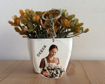 May include: A round clear keyring with a photo of a person holding a bouquet of flowers. The keyring has the name "Derek" printed on it.