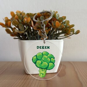 Puede incluir: Un llavero redondo de acrílico transparente con un brócoli verde de dibujos animados y el nombre "Derek" impreso en texto verde.