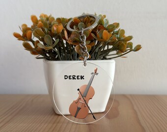 Puede incluir: Un llavero redondo de acrílico transparente con un violín marrón y el nombre "DEREK" impreso en negro.
