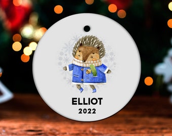 May include: A round white Christmas ornament with a cartoon hedgehog wearing a blue coat and holding a pine cone. The ornament has the name "ELLIOT" and the year "2022" printed on it.