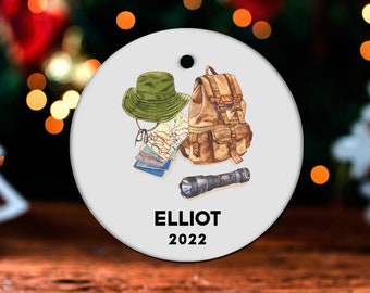May include: A white round Christmas ornament with a brown backpack, a green hat, a map, a flashlight, and the text "ELLIOT 2022".
