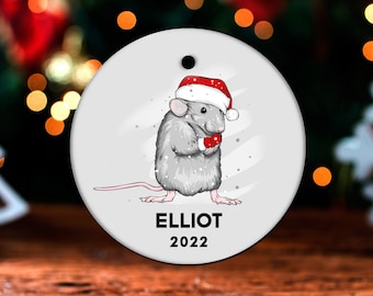 May include: A white round Christmas ornament with a gray mouse wearing a red Santa hat and holding a red mitten. The mouse is standing on a snowy background. The ornament has the name "ELLIOT" and the year "2022" printed on it.
