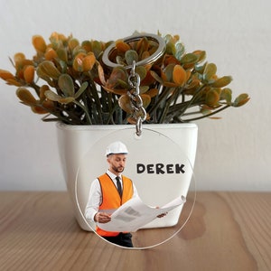 Puede incluir: Un llavero redondo y transparente con un llavero plateado. El llavero presenta un gráfico de una persona con casco blanco y chaleco naranja, sosteniendo un plano. El nombre "DEREK" está impreso en negro. El llavero se muestra frente a una planta en maceta.