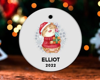May include: A round white Christmas ornament with a cartoon bear wearing a red Santa hat and holding a stack of presents. The ornament is decorated with snowflakes and the text "ELLIOT 2022".