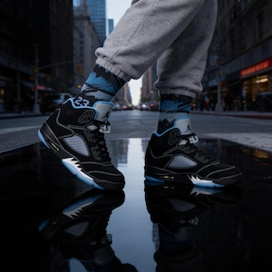 Puede incluir: Zapatillas deportivas de caña alta en ante negro con detalles en azul claro, con un panel lateral de malla, un cierre de cordones y una entresuela blanca con un diseño negro dentado. El logotipo Jumpman y el número 23 son visibles en el calzado.
