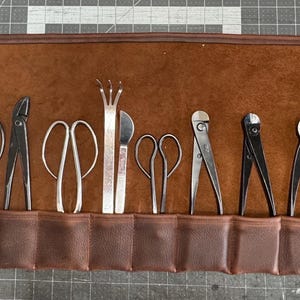 May include: A brown leather roll with 12 slots for holding gardening tools. The tools include pruning shears, a grafting tool, a serrated knife, and a pair of tweezers.