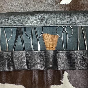 May include: A black leather tool roll featuring multiple individual pockets containing a variety of metal tools, including several pairs of scissors, pliers, tweezers, and a small brown brush, all organized in a horizontal row within the leather case.