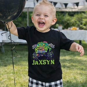 May include: A young child wearing a black t-shirt with a monster truck graphic and the name "Jaxsyn" in green. The child is holding a black balloon and smiling. The shirt features a green monster truck with a zombie-like face.