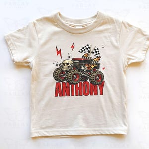 May include: Cream-colored t-shirt featuring a monster truck graphic with a skull, flames, and checkered flags. The name "ANTHONY" is printed in red block letters below the truck. The truck has large tires and red accents.