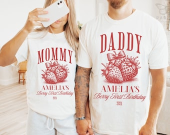 May include: Matching off-white t-shirts with red text and strawberry graphics. One shirt says "MOMMY" and the other says "DADDY". Both shirts include "AMELIA'S Berry First Birthday 2024".