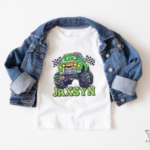 Puede incluir: Camiseta blanca con un gr&aacute;fico de cami&oacute;n monstruo verde y el nombre "JAXSYN" en verde. El cami&oacute;n monstruo tiene un tema zombi con cerebros expuestos y una bandera a cuadros. Una chaqueta vaquera azul est&aacute; sobre la camiseta.