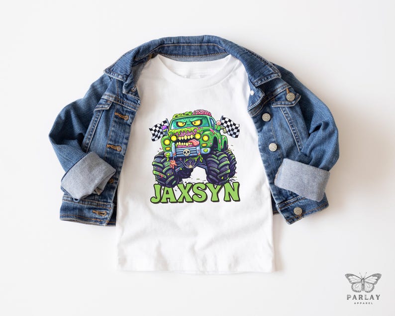 Puede incluir: Camiseta blanca con un gr&aacute;fico de cami&oacute;n monstruo verde y el nombre "JAXSYN" en verde. El cami&oacute;n monstruo tiene un tema zombi con cerebros expuestos y una bandera a cuadros. Una chaqueta vaquera azul est&aacute; sobre la camiseta.
