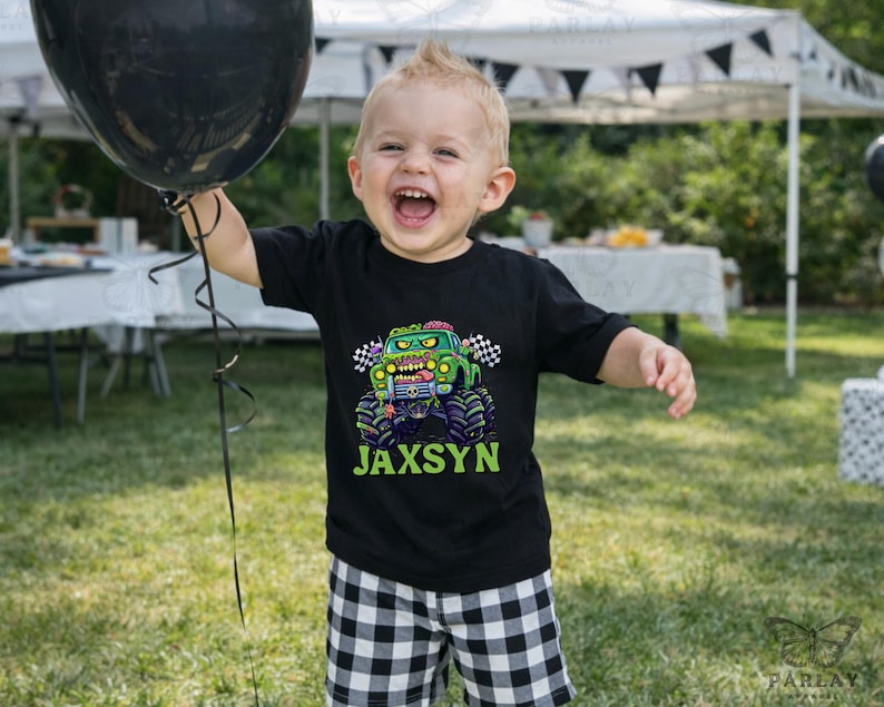 May include: A young child wearing a black t-shirt with a monster truck graphic and the name "Jaxsyn" in green. The child is holding a black balloon and smiling. The shirt features a green monster truck with a zombie-like face.