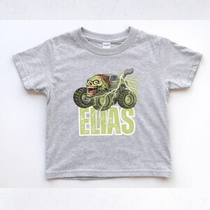 May include: A light gray toddler t-shirt featuring a monster truck with a zombie driver and the name "ELIAS" in green block letters. The truck has large tires and is surrounded by lightning bolts. The shirt is made of a soft material.