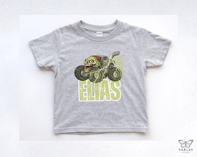 May include: A light gray toddler t-shirt featuring a monster truck with a zombie driver and the name "ELIAS" in green block letters. The truck has large tires and is surrounded by lightning bolts. The shirt is made of a soft material.