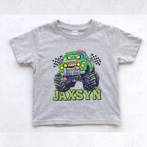 Pu&ograve; includere: Una maglietta grigia a maniche corte con un monster truck verde con un design a tema zombie. Il camion ha pneumatici grandi, una bandiera a scacchi e il nome "JAXSYN" in lettere verdi. La maglietta &egrave; realizzata in materiale morbido.