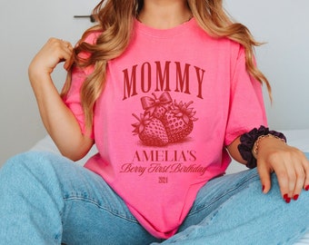 May include: Pink t-shirt with the text "Mommy Amelia's Berry First Birthday 2024" in red lettering. The shirt features a graphic of two strawberries with a bow.