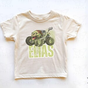 May include: A cream-colored t-shirt featuring a green monster truck with a zombie driver and the name "ELIAS" in green block letters. The graphic includes lightning bolts and smoke effects, creating a playful, cartoonish design.