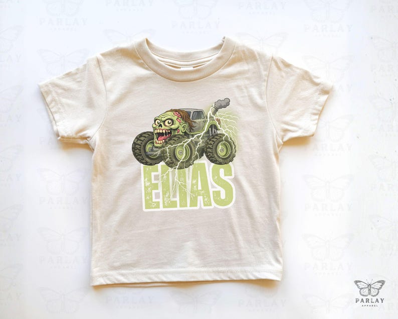 May include: A cream-colored t-shirt featuring a green monster truck with a zombie driver and the name "ELIAS" in green block letters. The graphic includes lightning bolts and smoke effects, creating a playful, cartoonish design.