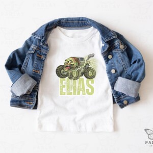May include: White t-shirt featuring a green zombie monster truck design with the name "ELIAS" in green block letters. A denim jacket is draped over the shirt. The design includes lightning bolts and a cartoon zombie face.