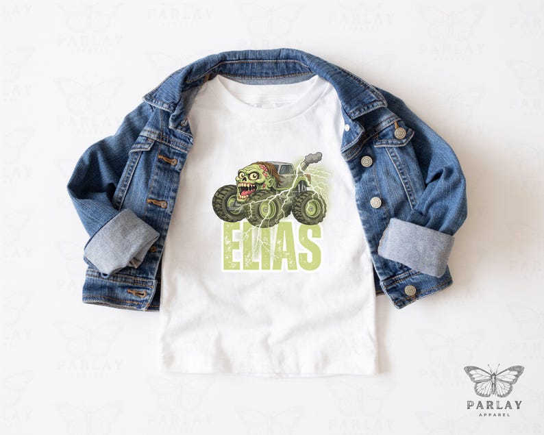 May include: White t-shirt featuring a green zombie monster truck design with the name "ELIAS" in green block letters. A denim jacket is draped over the shirt. The design includes lightning bolts and a cartoon zombie face.