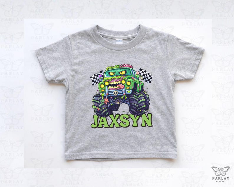 Puede incluir: Una camiseta gris de manga corta con un dise&ntilde;o de cami&oacute;n monstruo verde con tem&aacute;tica zombi. El cami&oacute;n tiene neum&aacute;ticos grandes, una bandera a cuadros y el nombre "JAXSYN" en letras verdes. La camiseta es de un material suave.