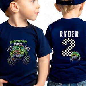 May include: Navy blue short-sleeved t-shirt featuring a monster truck design. The front displays "BIRTHDAY BOY" above a green monster truck with checkered flags. The back has "RYDER" and the number "2".