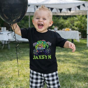 Puede incluir: Un ni&ntilde;o lleva una camiseta negra con un dise&ntilde;o de cami&oacute;n monstruo y el nombre "Jaxsyn" en verde. El ni&ntilde;o sostiene un globo negro y sonr&iacute;e. El fondo incluye una carpa blanca y hierba verde.