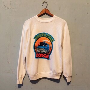 Op de afbeelding: Wit sweatshirt met een afbeelding van een blauwe bestelwagen met de tekst "VMTASIC DODGE" in een cirkel.