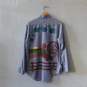 May include: A pale blue button-down shirt with a painted farm scene on the back. The design features a rooster, a sunrise, a fence, and blue floral accents. The shirt is hanging on a wooden hanger.