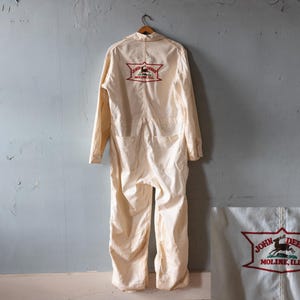 Op de afbeelding: Crèmekleurige vintage jumpsuit met lange mouwen en zakken. De achterkant heeft een rood en groen geborduurd logo met de tekst "JOHN DEERE MOLINE, ILL." De jumpsuit hangt aan een houten kleerhanger.