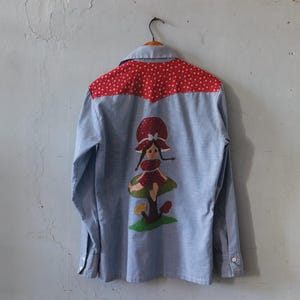 May include: A light blue button-down shirt with a red polka-dot yoke. The back features an embroidered design of a girl sitting on a mushroom. The girl is wearing a red dress and a mushroom hat.