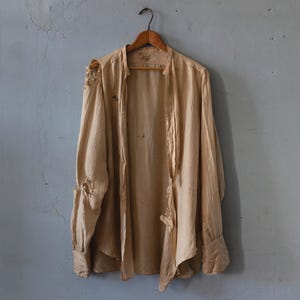 Op de afbeelding: Een vintage beige shirt met lange mouwen hangt aan een houten hanger. Het shirt heeft een relaxte pasvorm en lijkt gemaakt van een lichte stof. Het kledingstuk vertoont tekenen van slijtage, wat bijdraagt aan de antieke uitstraling.