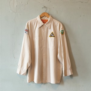 May include: A striped, beige long-sleeved shirt with three patches. The patches are a red and blue rectangle, a green triangle with a white logo, and a green circle with a white logo.