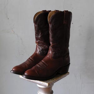 Könnte beinhalten: Ein Paar braune Leder-Cowboystiefel. Die Stiefel haben eine spitze Zehenpartie, dekorative Nähte und einen hohen Schaft. Die Stiefel werden auf einem kleinen, dekorativen Ständer ausgestellt. Die Stiefel scheinen Vintage zu sein.