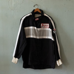 Könnte beinhalten: Schwarze Rennjacke mit weißen Streifen auf den Ärmeln und einem grauen Streifen über der Brust. Die Jacke trägt den Text "Safety Racing" auf der Brust und einen kleinen Aufnäher auf dem Ärmel.