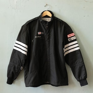 May include: Black racing jacket with white stripes on the sleeves. Includes patches with the "Simpson" logo and an American flag. The jacket has a zip-up front and the name "Mac Evans" is embroidered on the chest.