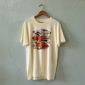 Puede incluir: Camiseta color crema con un gráfico de tres coches clásicos en morado, rojo y naranja. El diseño incluye el texto "HEMI" y "Rapid Transit 500" en un estilo retro. La camiseta está colgada de una percha de madera.